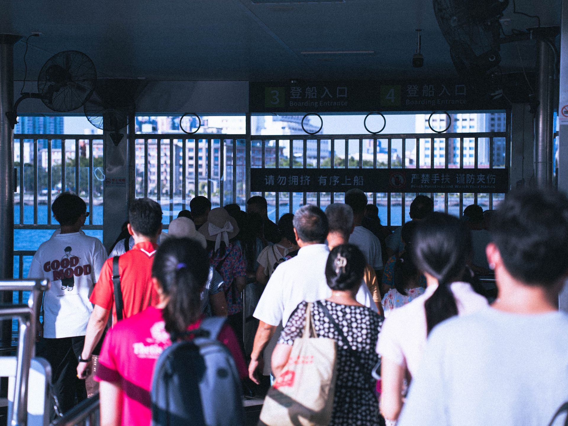 A diverse crowd waits to board at a waterfront ferry terminal on a sunny day.