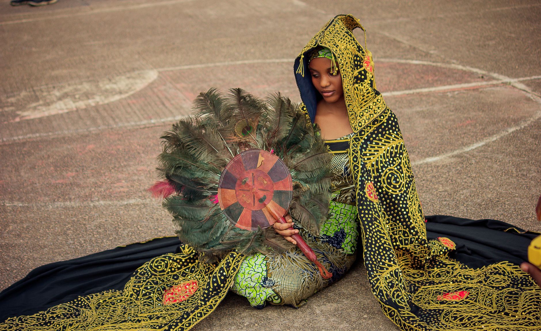 A woman in vibrant African attire sits with a decorative feather fan, showcasing cultural elegance.