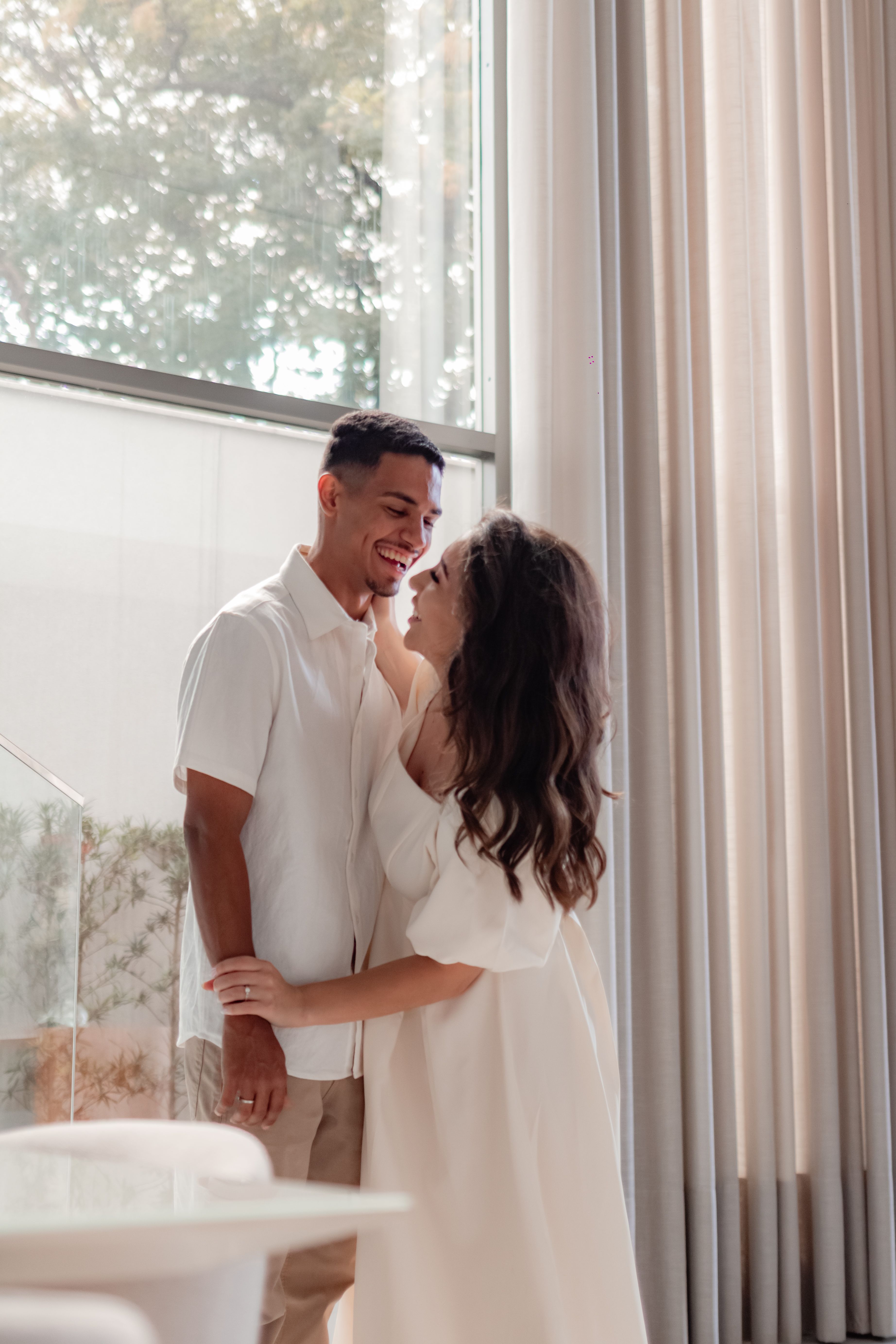 A joyful couple in white clothing embraces by a large window, radiating warmth and love indoors.