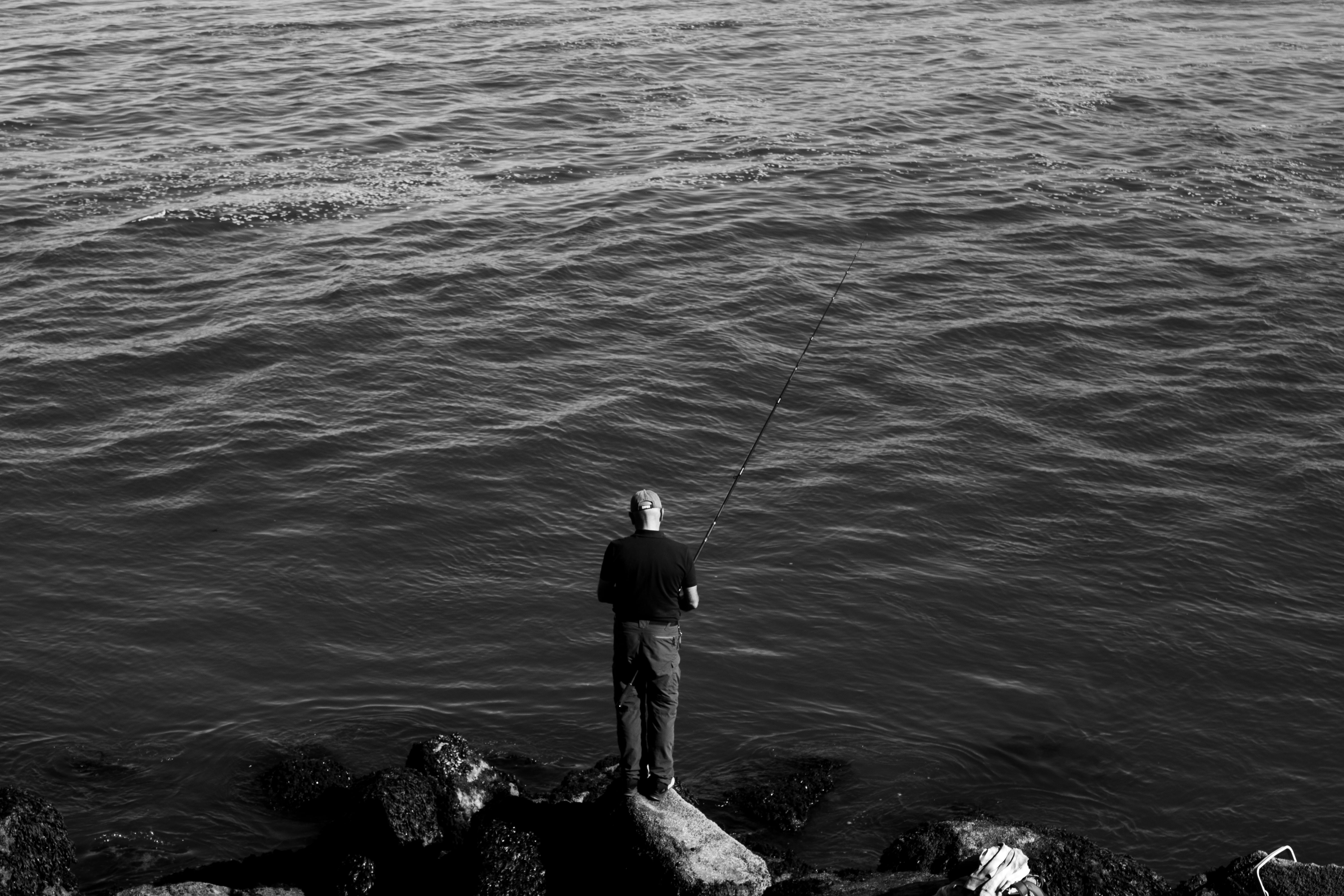 在海边岩石上，一名男子正专注地钓鱼，形成一幅黑白剪影的画面