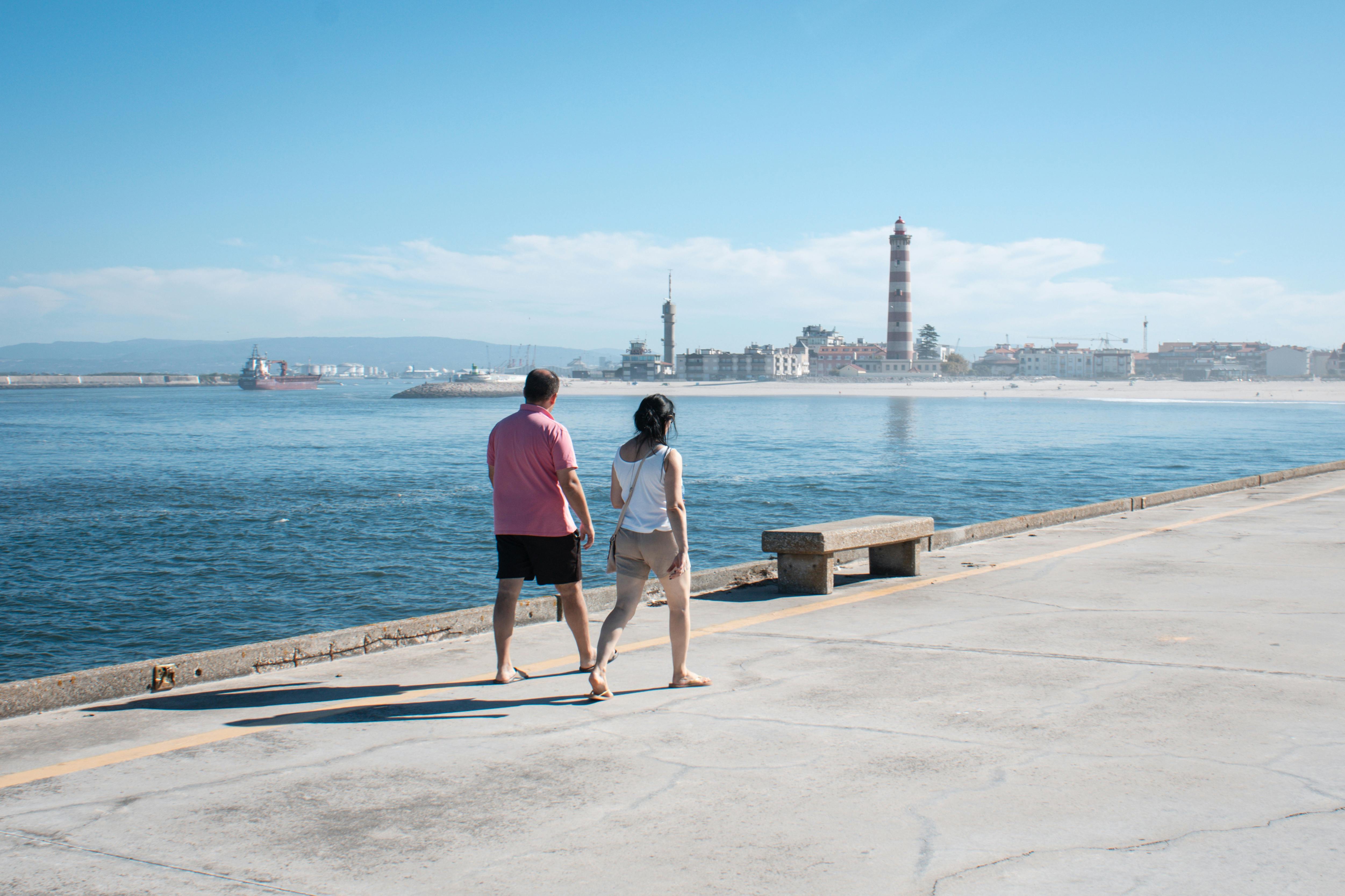 一对夫妇沿着海滨散步，远处的灯塔和清澈的蓝天