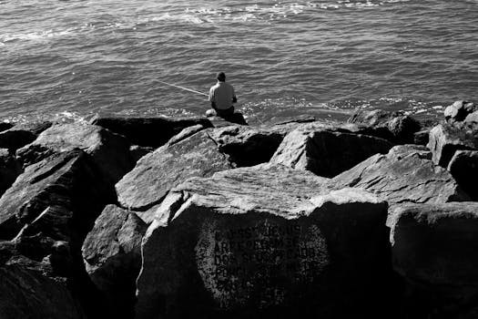 一个孤独的渔夫坐在海边的岩石上，在黑白的场景中投下他的线