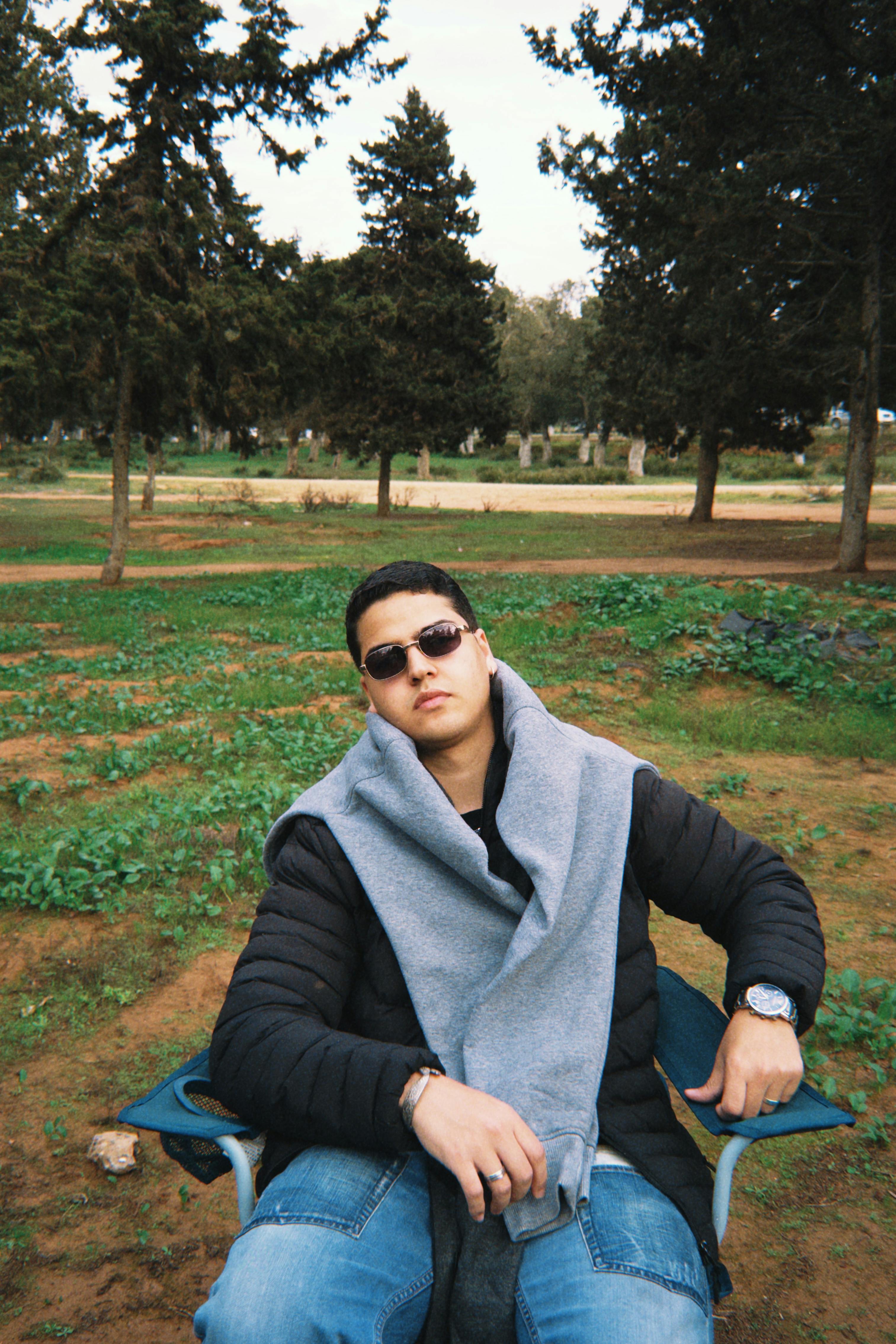 Young man in sunglasses relaxing in a park setting with trees in the background.