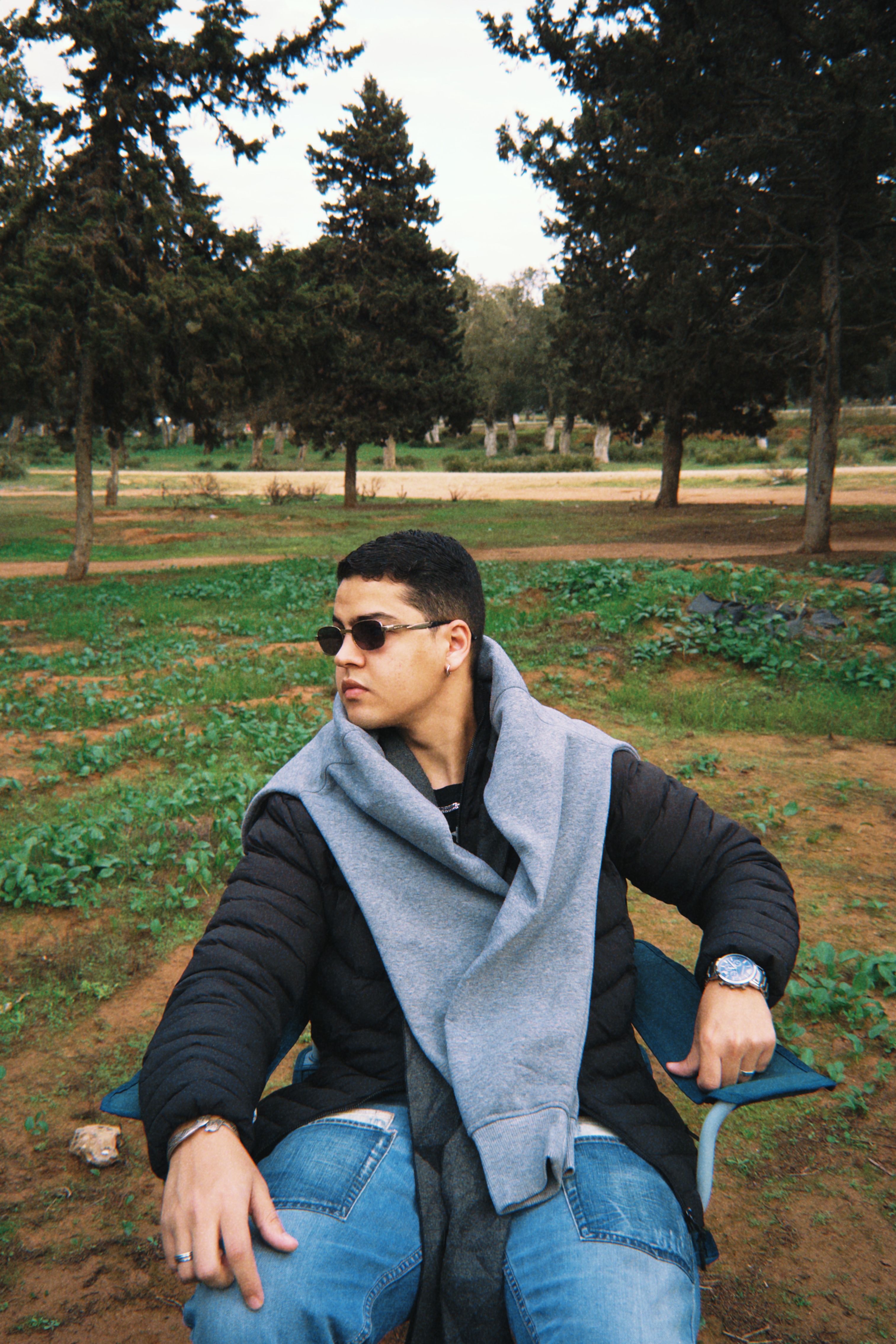Stylish young man sitting outdoors in a wooded park, wearing sunglasses and casual attire.