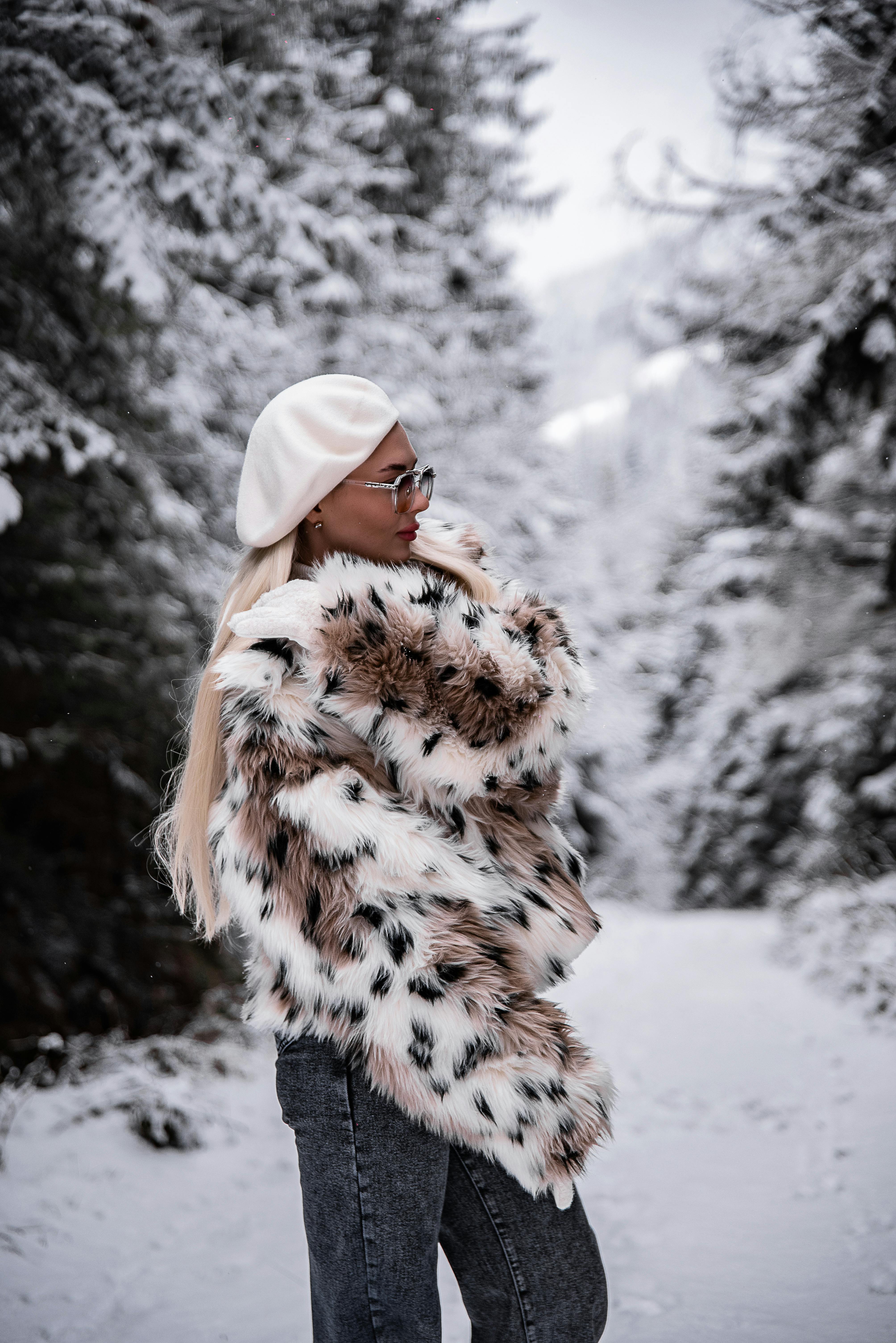 穿着人造毛皮夹克，戴着贝雷帽的时尚女子在白雪皑皑的森林小路上摆姿势