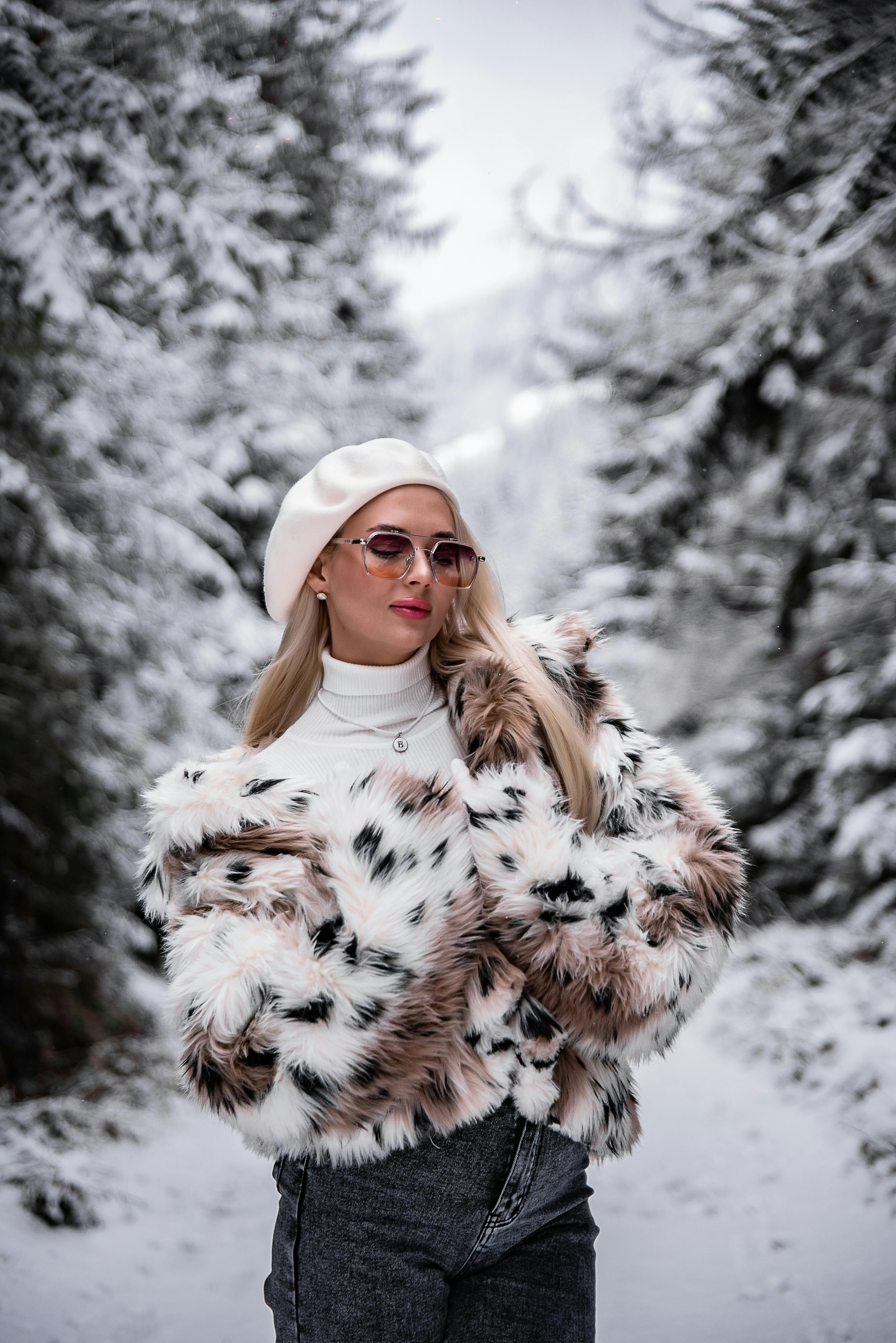 在白雪覆盖的森林景致中，一位穿着人造毛皮外套、戴着贝雷帽的时尚女子正摆出姿势