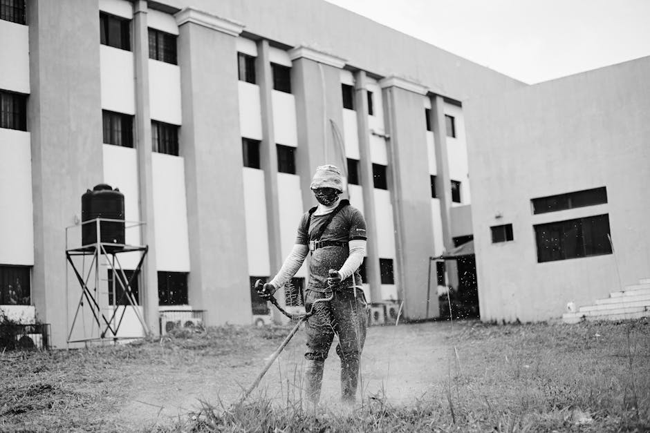 A masked worker trims grass with machinery outside a large building.