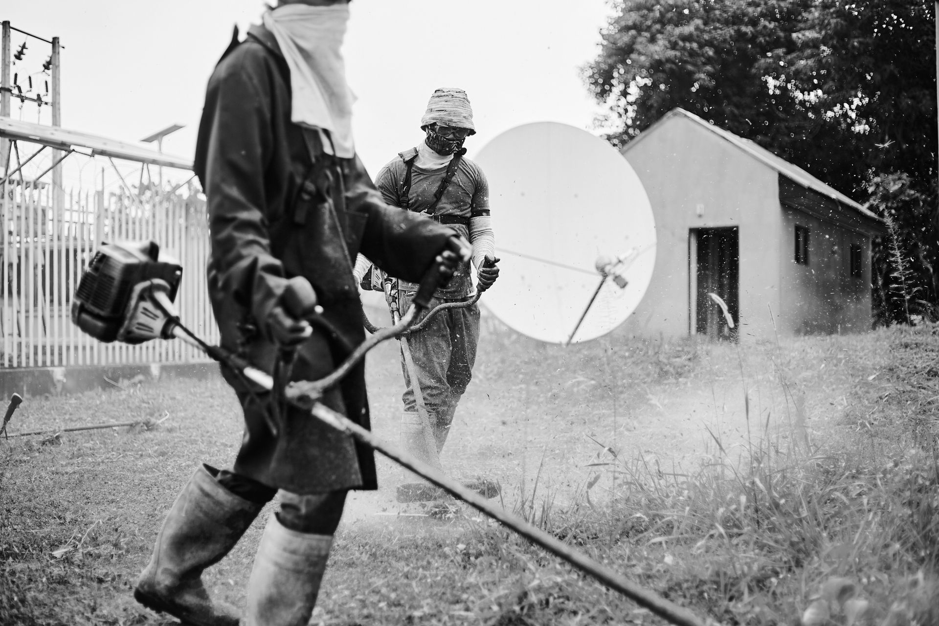 Two workers trimming grass outside an industrial facility with protective gear.