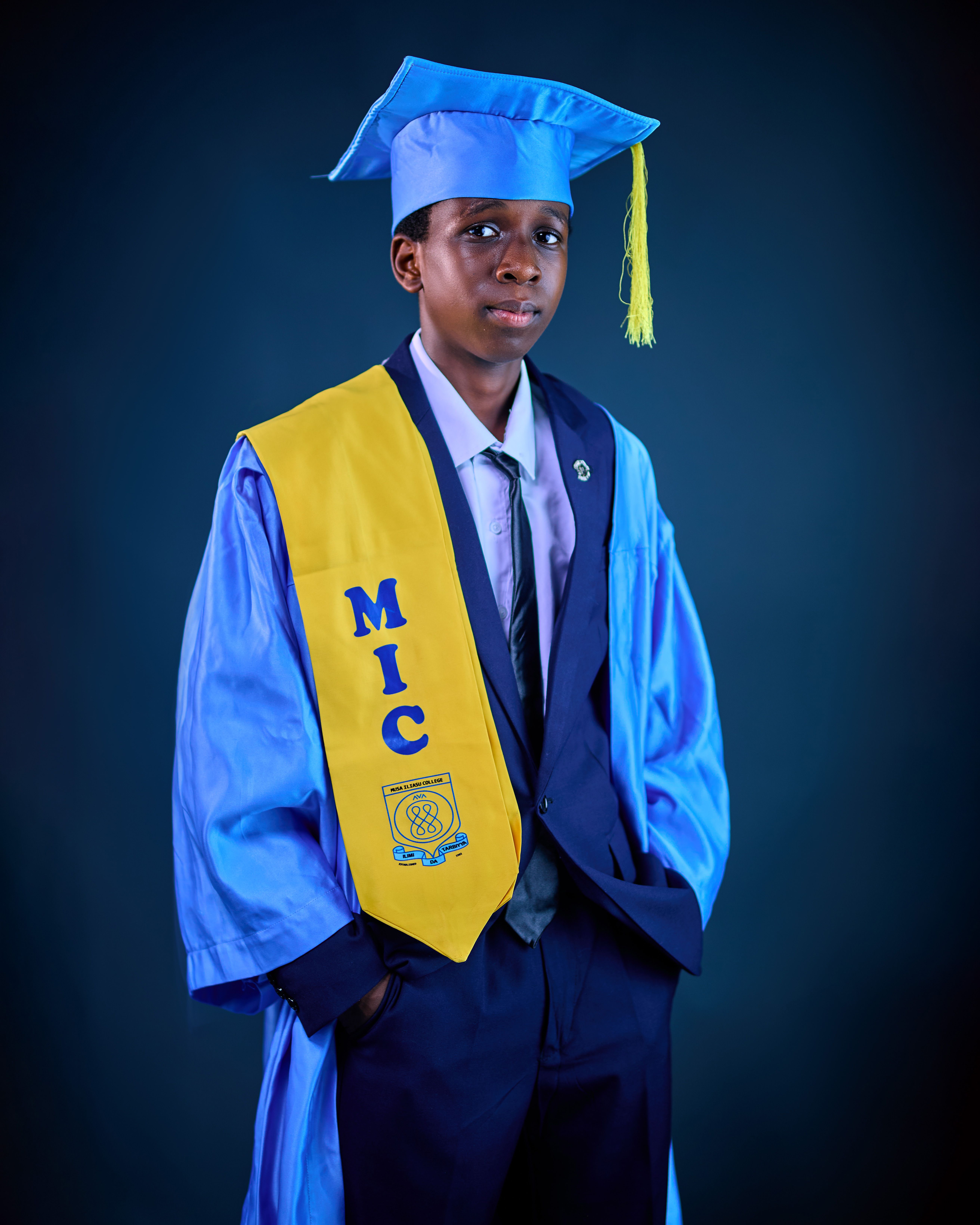 Young male graduate in blue cap and gown with yellow stole on dark background.