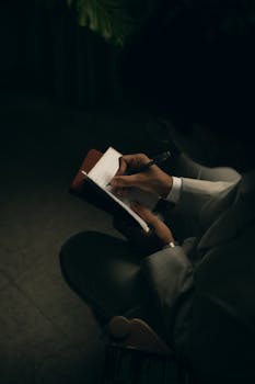 An individual writing in a notebook in a dimly lit setting, capturing a reflective moment.
