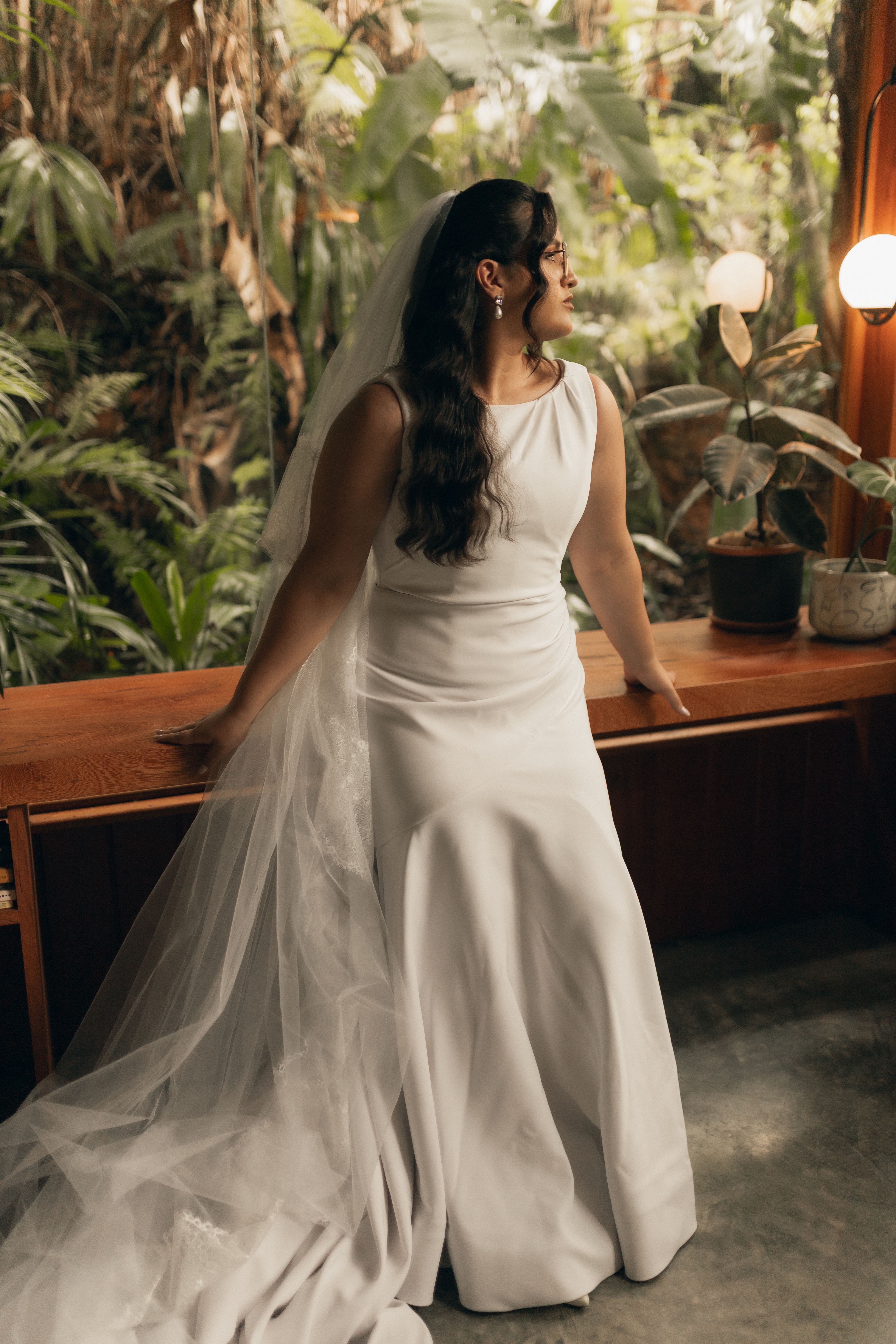 Bride in a classic white dress indoors with a lush plant backdrop.