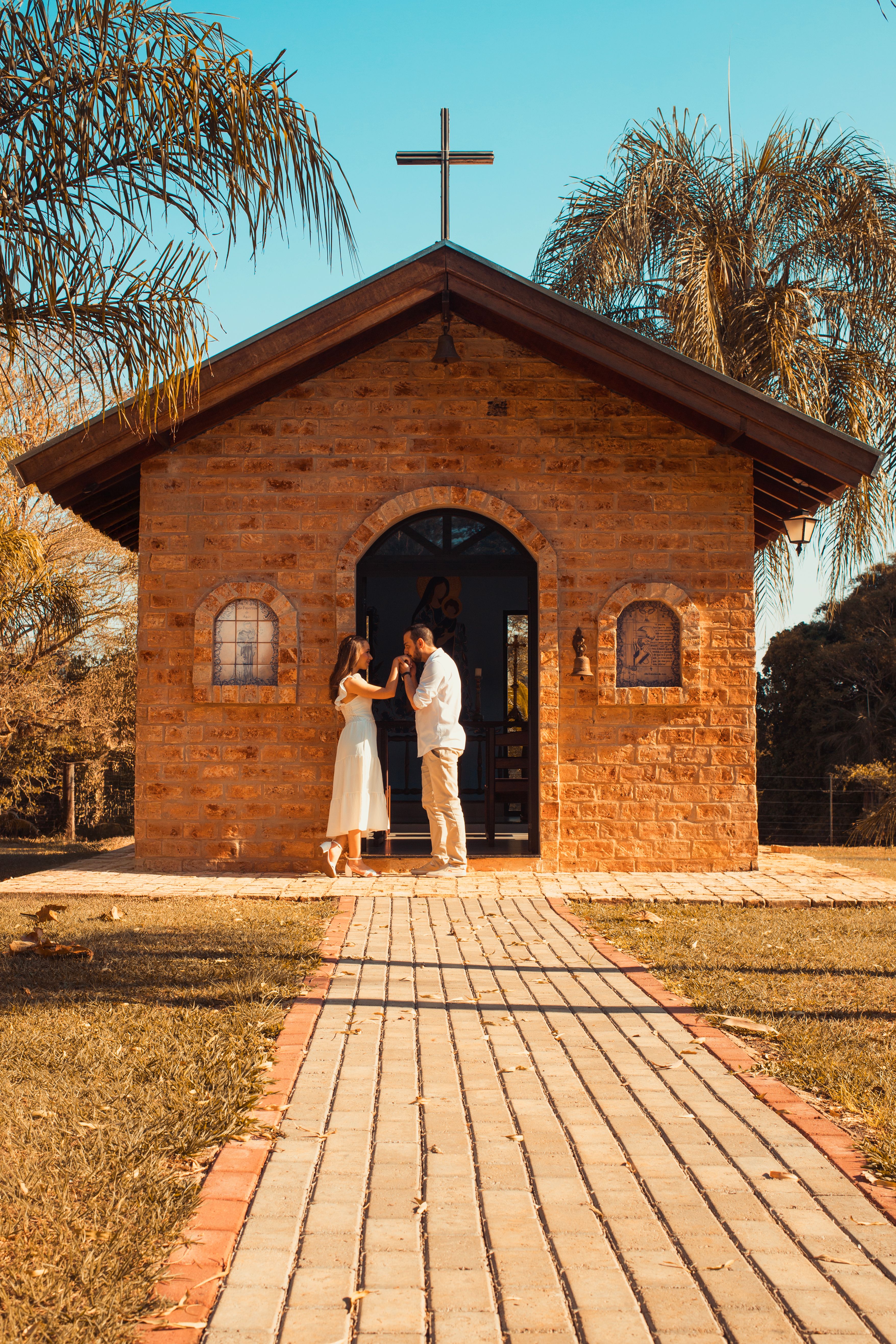 A couple in front of a rustic brick chapel under palm trees during sunset.