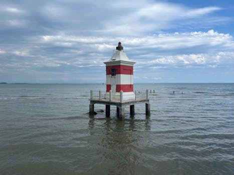 红白相间的灯塔矗立在平静的蓝色大海中，其头顶散布着朵朵云彩