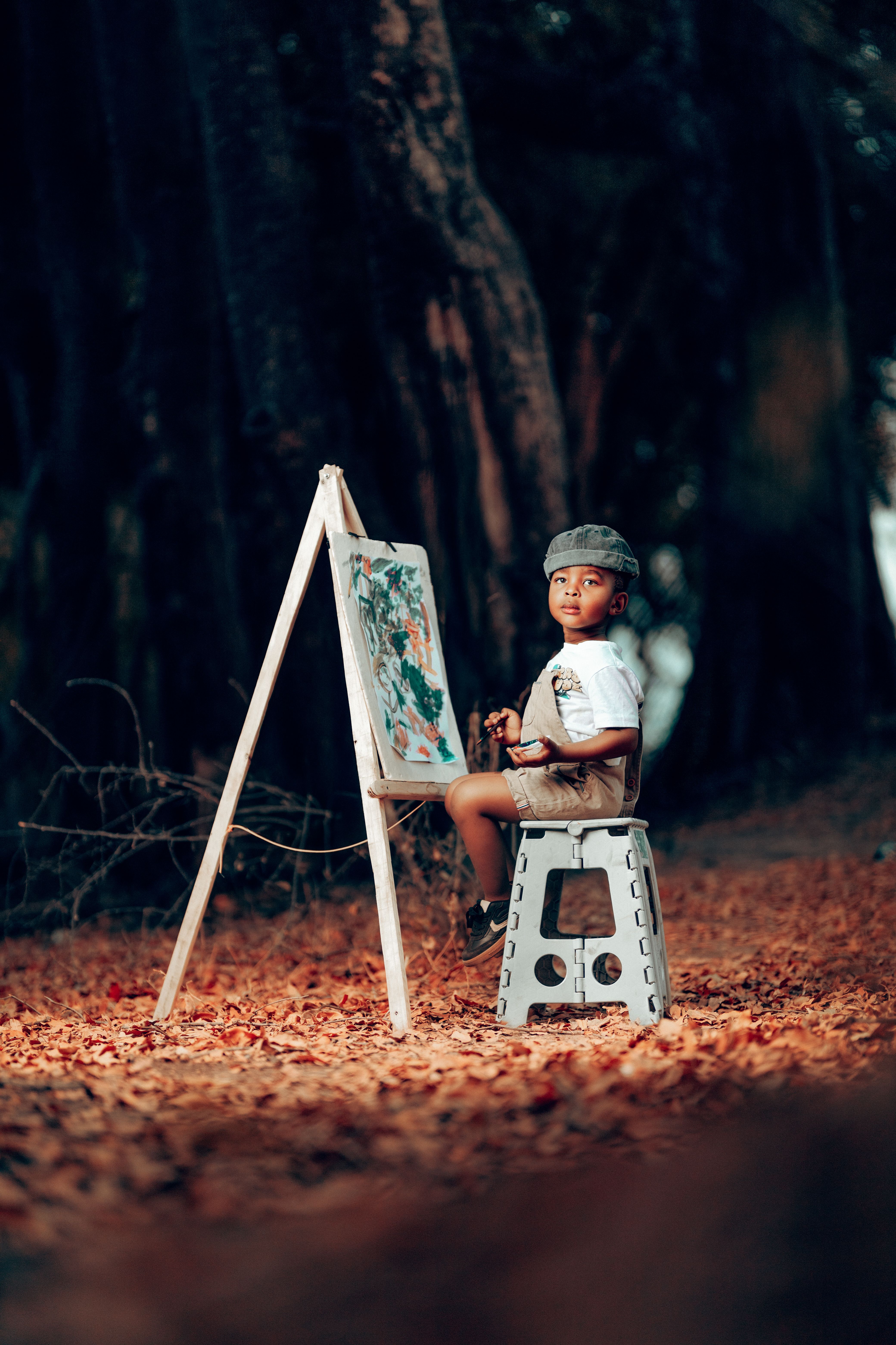 Child sitting on a stool painting outdoors amid autumn leaves.