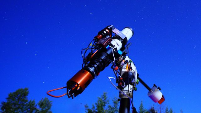 An imaging telescope with cameras and cables on an equatorial mount under a clear blue evening sky