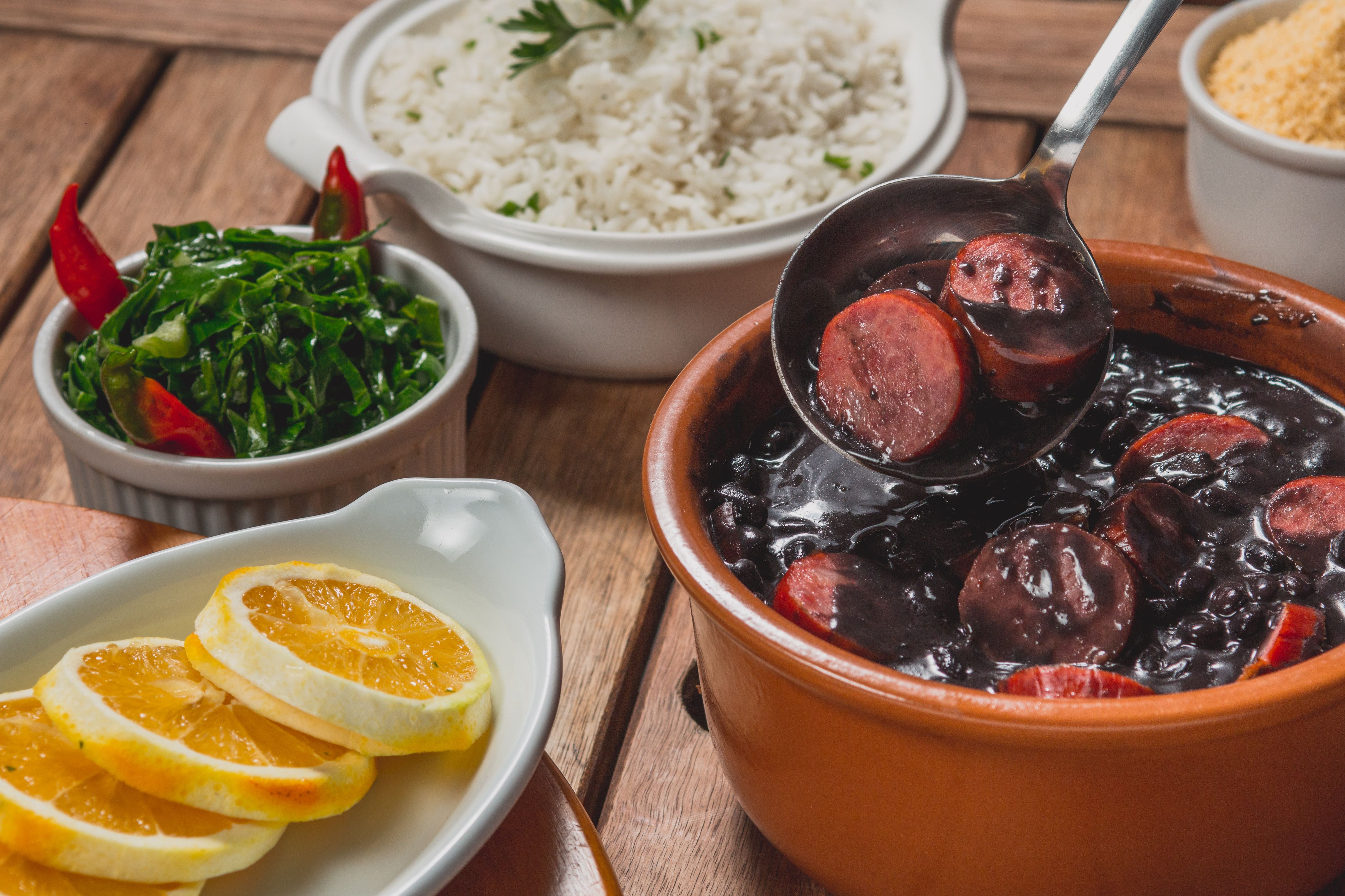Feijoada na panela de barro com arroz, couve e laranja na mesa de madeira