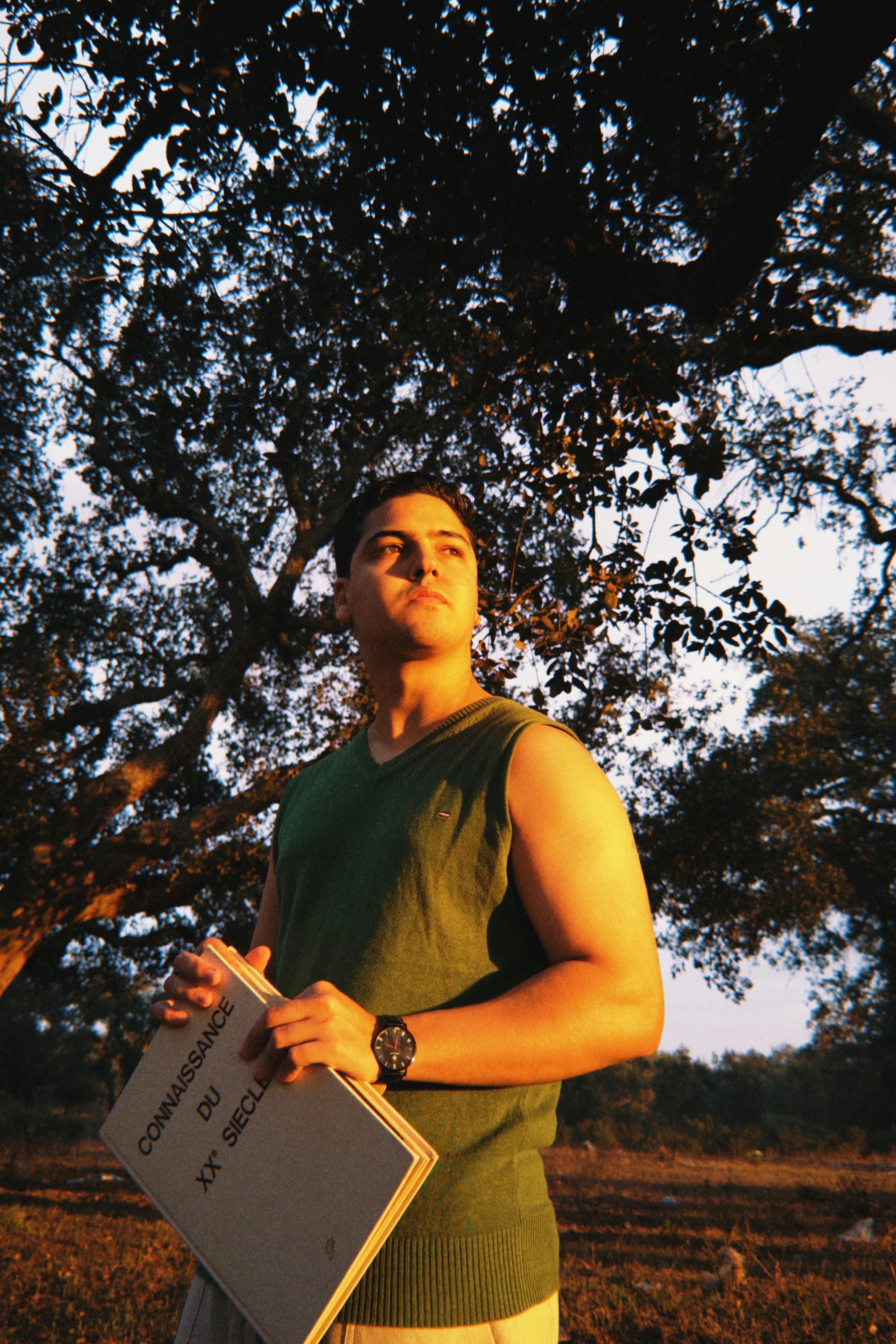 Young man in green vest holding a book outdoors during sunset under a tree, exuding confidence.