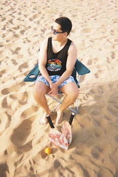 一个年轻人坐在沙滩椅上，阳光明媚的天空下，沙滩上放着桨球