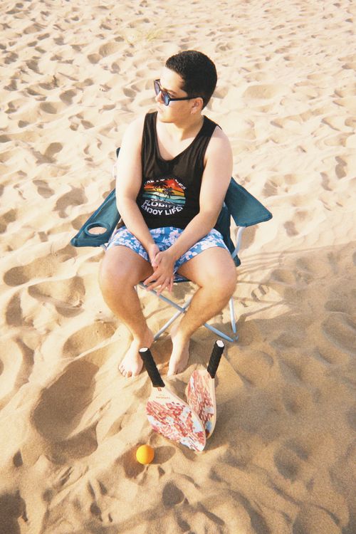 Free A young man sitting on a beach chair with paddle ball set in the sand under sunny skies. Stock Photo