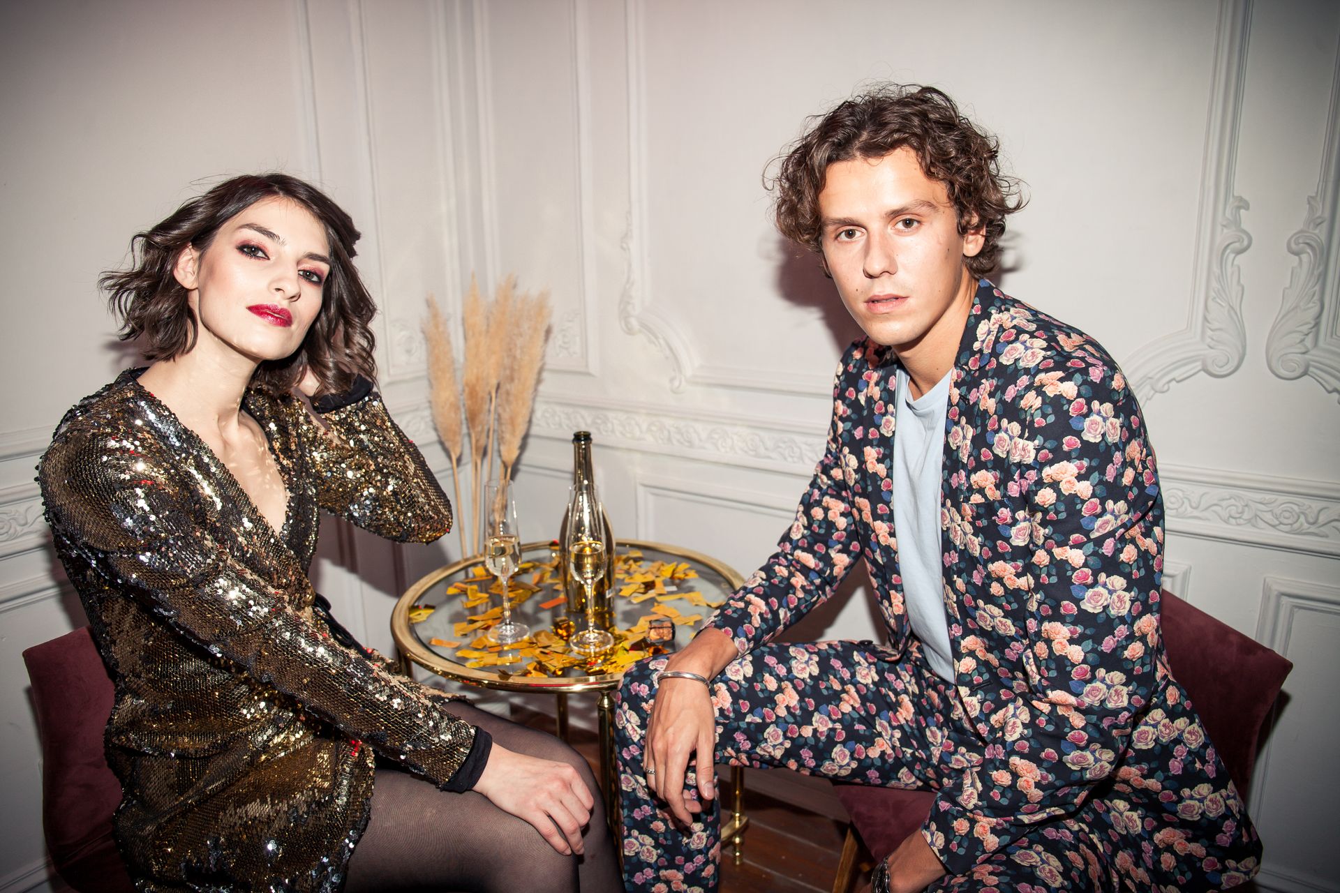 Elegant couple in fashionable attire enjoying drinks at an indoor celebration.