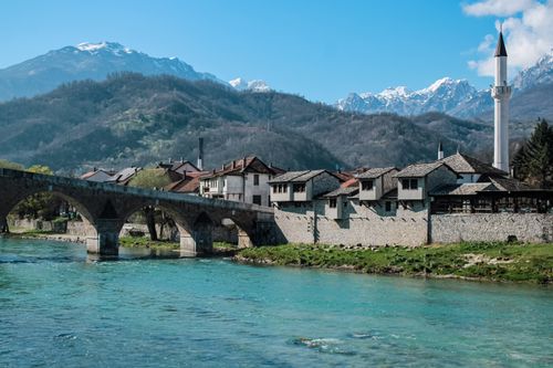 Free Picturesque stone bridge and mosque set against stunning mountain backdrop with clear blue river. Stock Photo
