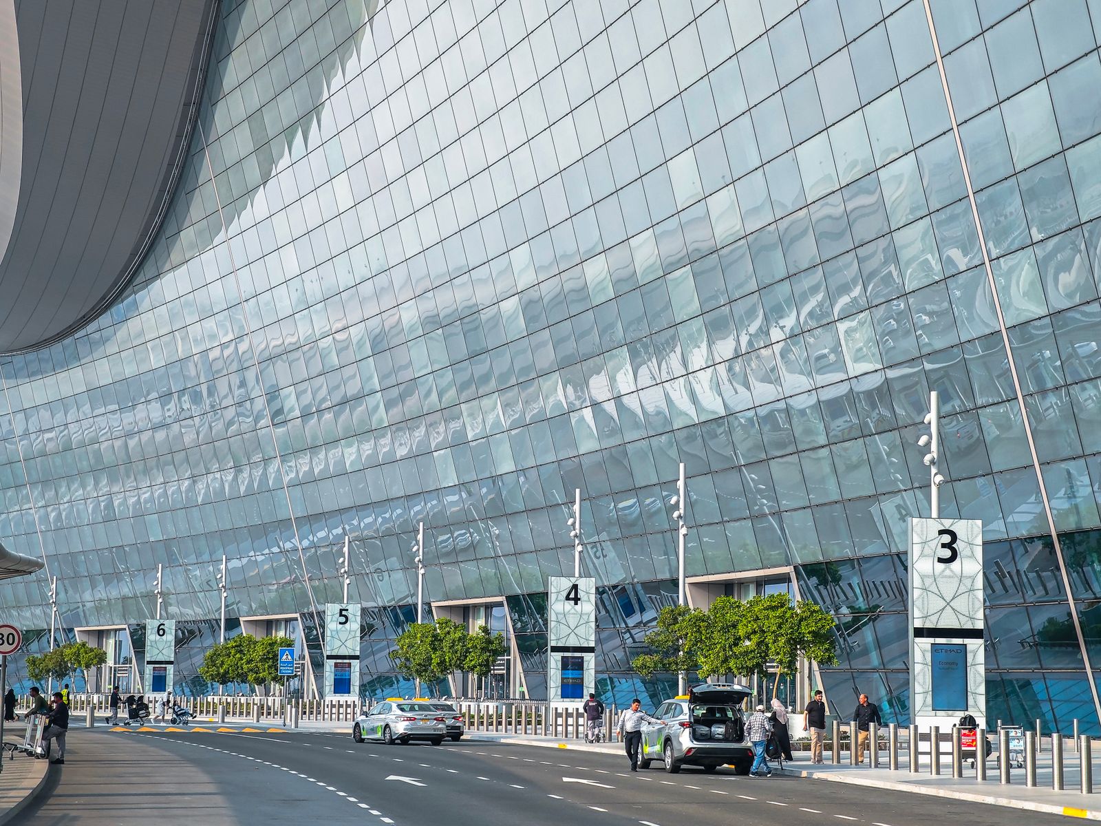 Kerbside drop-off lanes beside the curved glass facade of an Abu Dhabi airport terminal