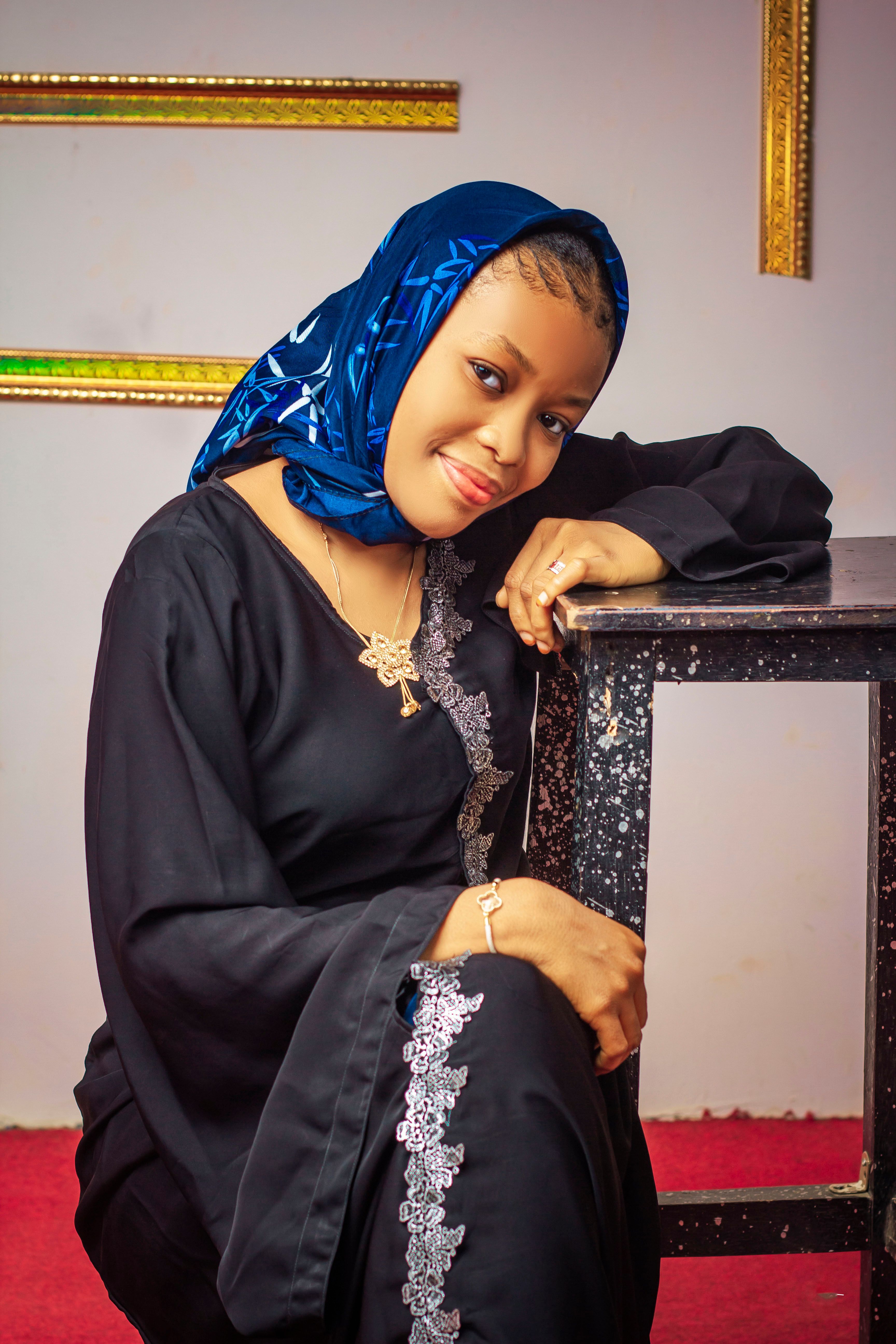 Smiling woman in traditional black dress and blue headscarf posing indoors, exuding elegance.