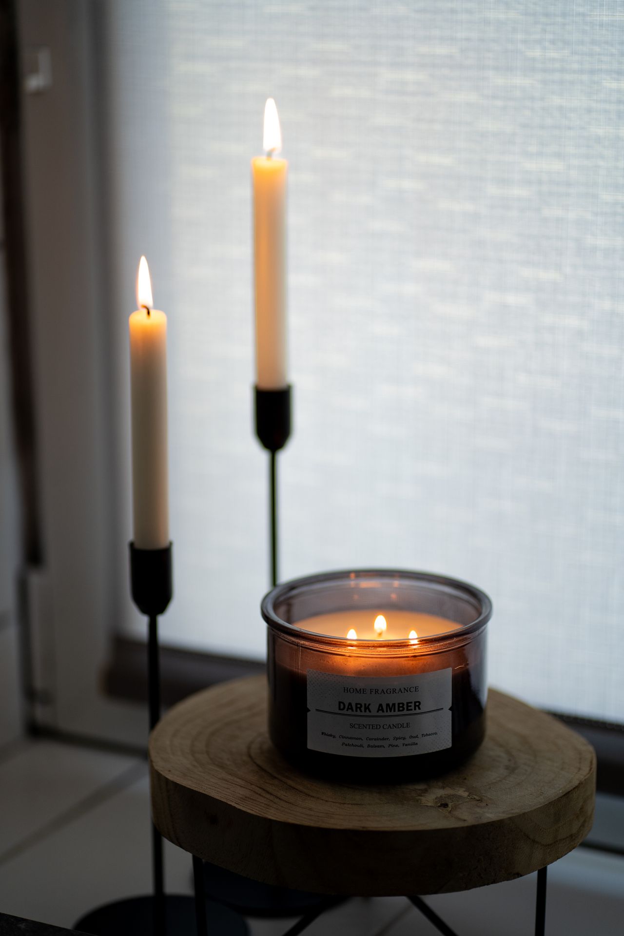 Dark amber scented candle with lit taper candles in black holders on a wooden table