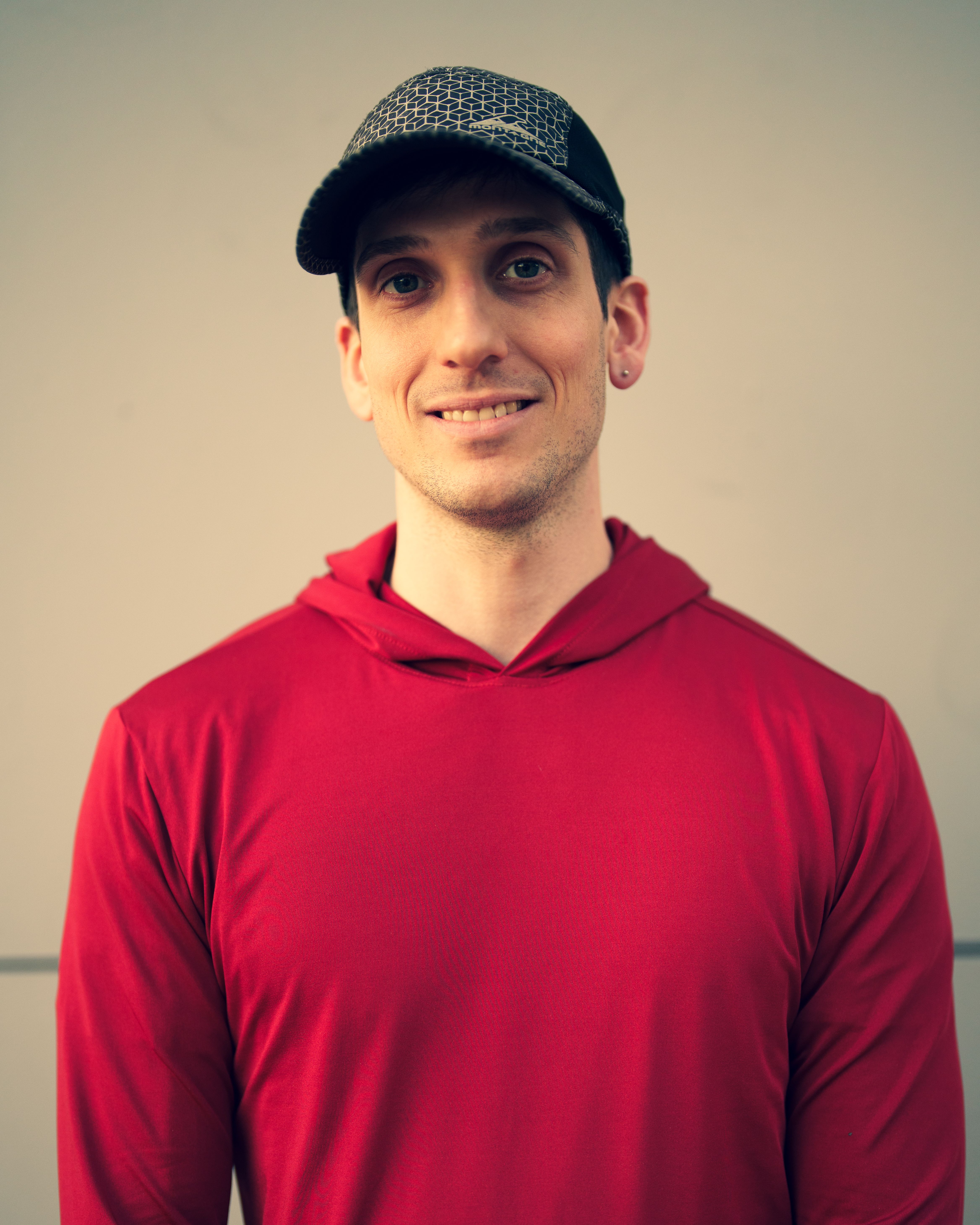 Portrait of a man in a red hoodie and cap standing against a plain wall.