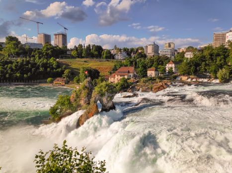 Explore the powerful Rhine Falls in Neuhausen am Rheinfall, Switzerland, surrounded by vibrant greenery and urban architecture.