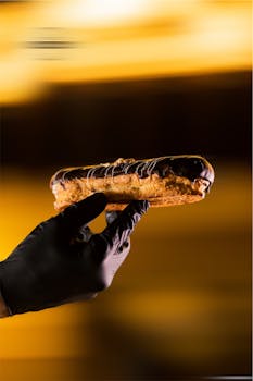 A gloved hand holds a delicious chocolate éclair with soft yellow and brown tones.