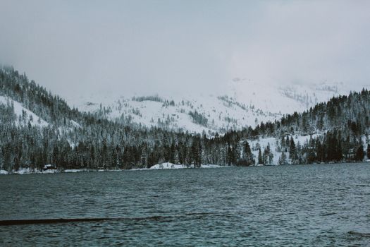 Snow-capped mountains and pine trees surround a tranquil lake in winter, offering a peaceful scenic view.