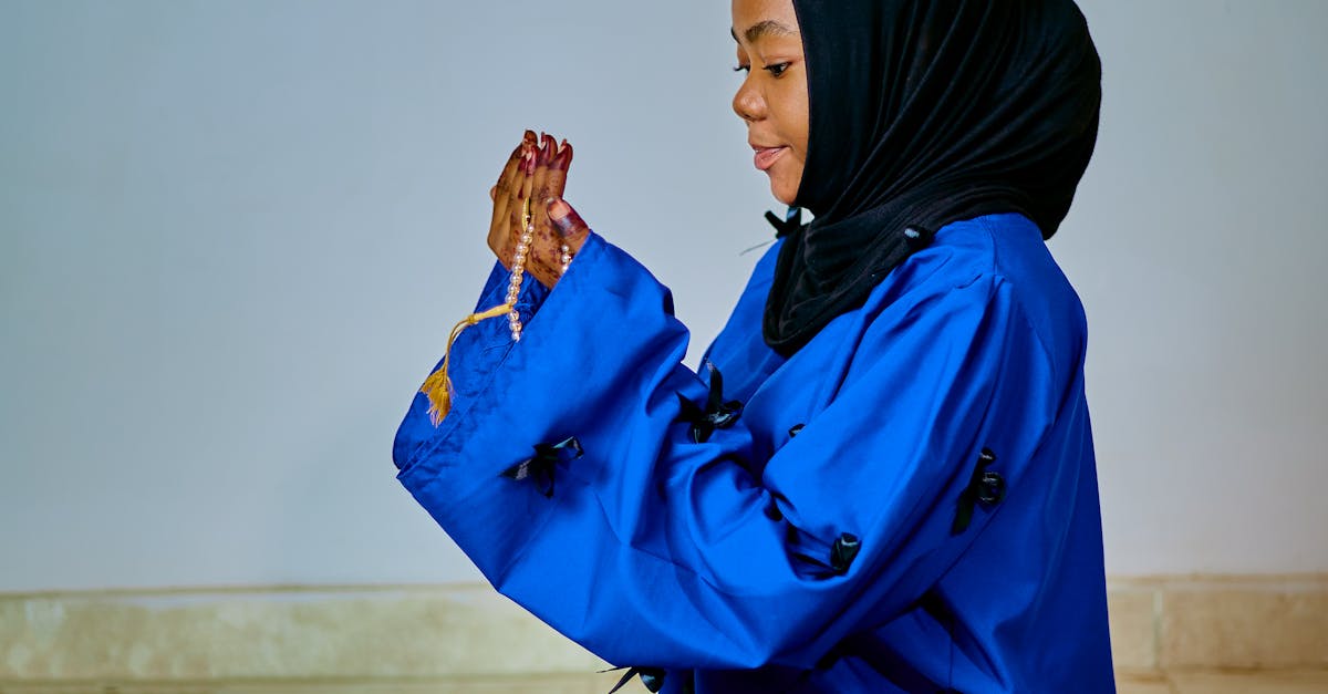 A young woman in a blue robe and hijab, sitting and praying with prayer beads indoors.