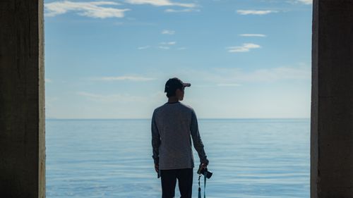 Free A solitary person holding a camera gazes out over the calm sea, framed by concrete pillars. Stock Photo