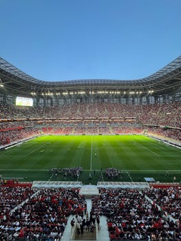 A lively soccer match unfolds before a packed stadium under a clear blue sky.