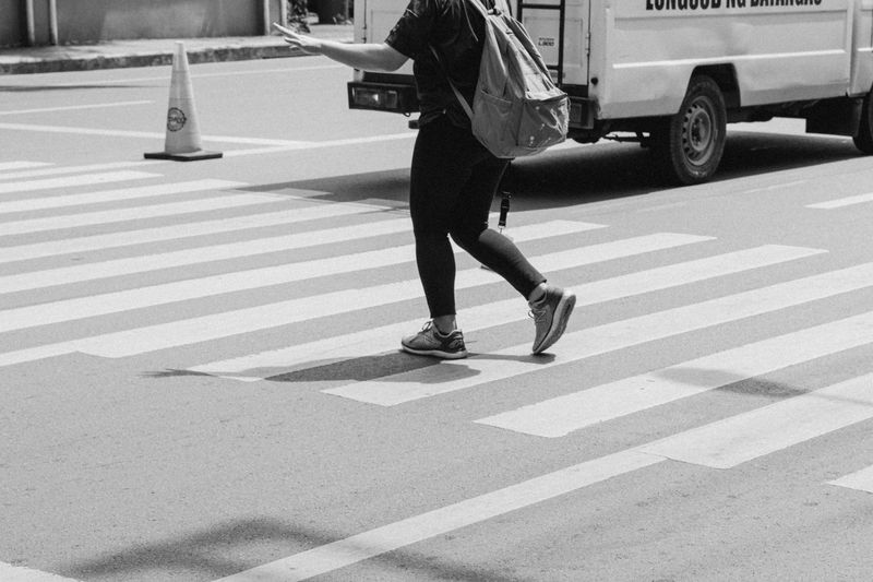 Black and white photo of a person crossing a city crosswalk with a backpack and a van nearby.