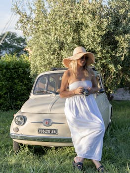 Lady in white dress and straw hat poses by a vintage Fiat car outdoors.