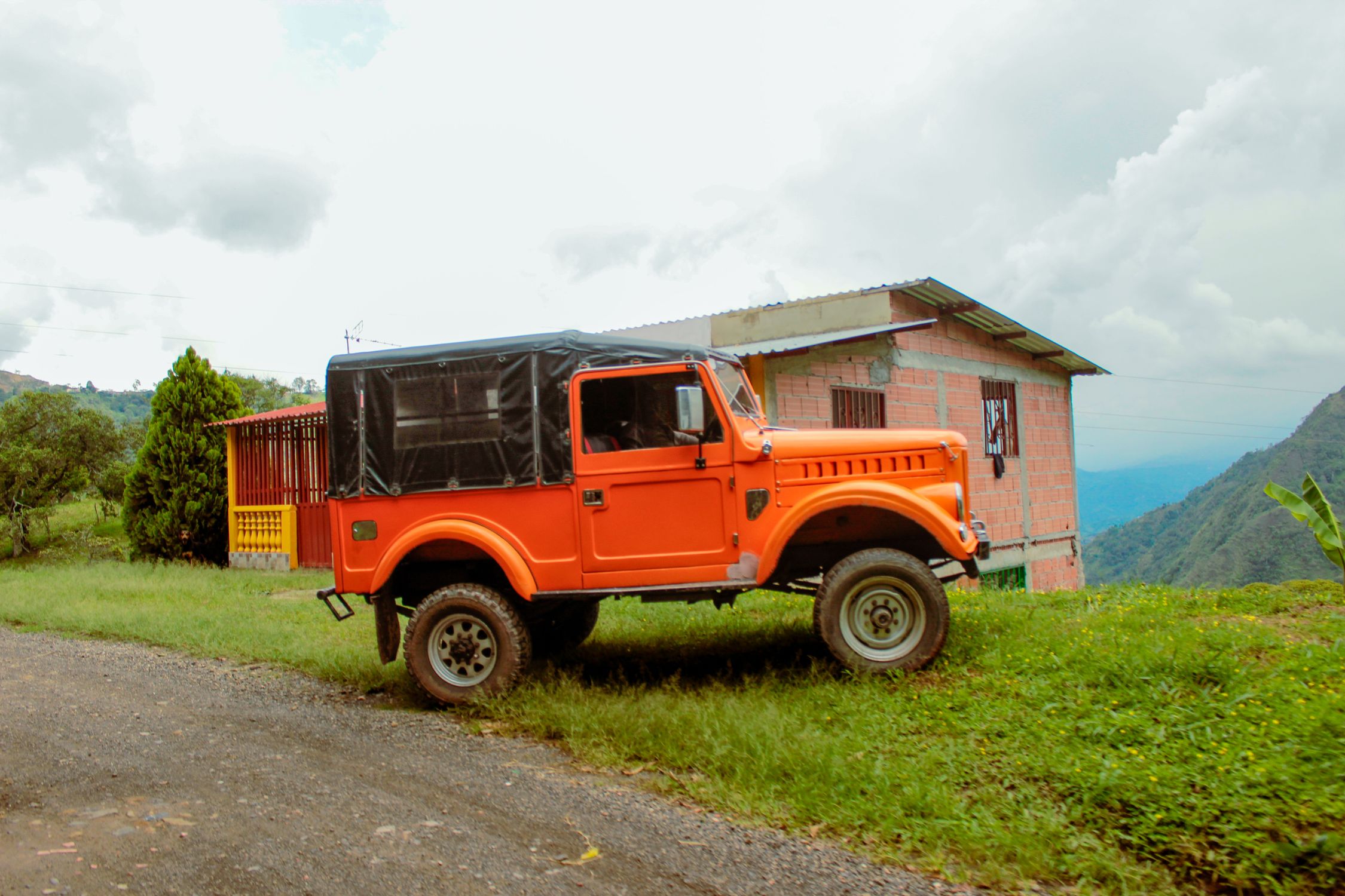 Bright orange off-road jeep parked near a rustic house in a lush green rural landscape.