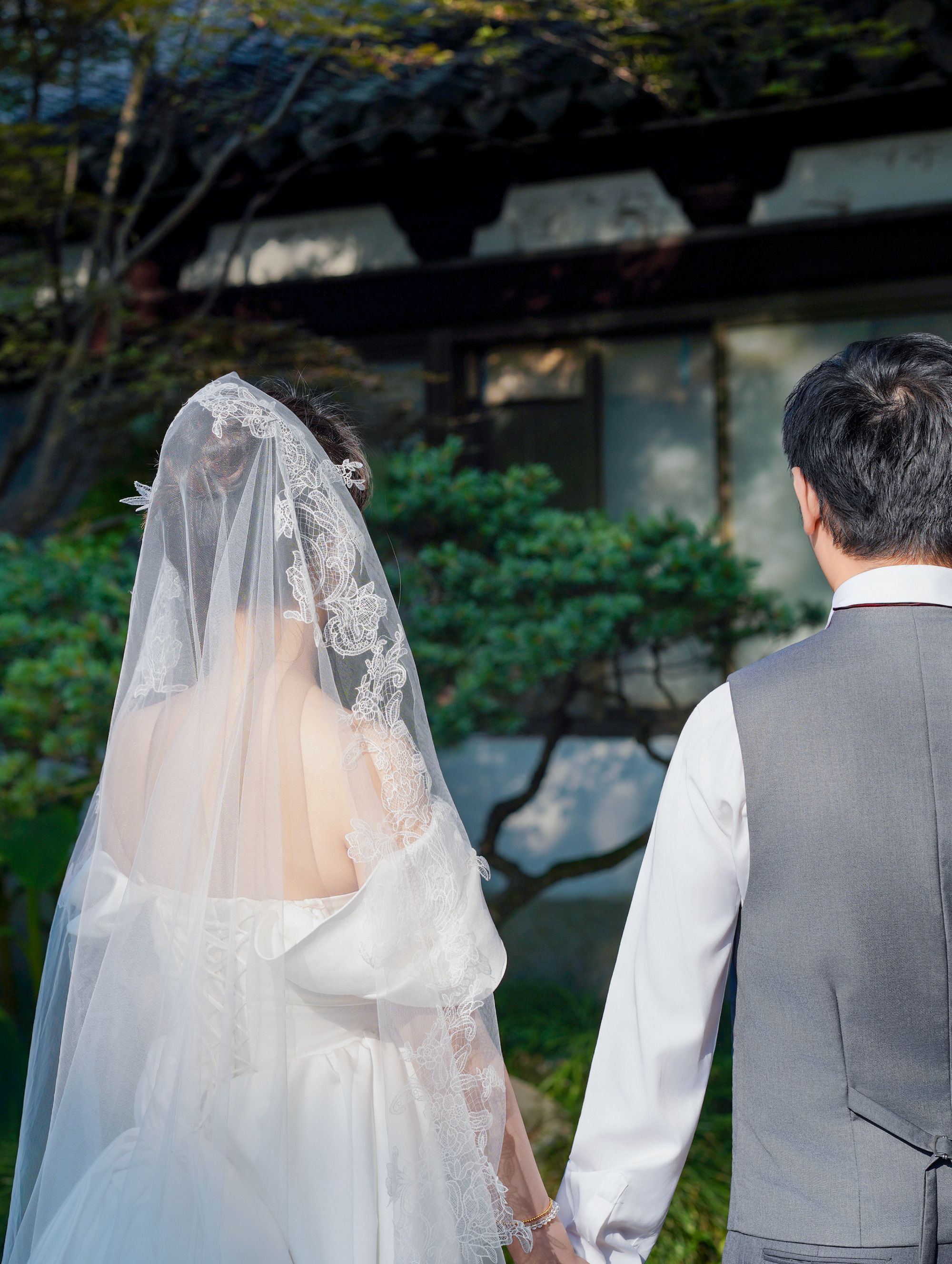 Bride and groom holding hands outdoors in a traditional setting, back view.