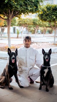 A man in white attire poses with two dogs in a sunny urban park setting.