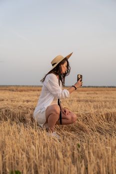 A woman in a straw hat takes a photo in a golden wheat field during sunset.