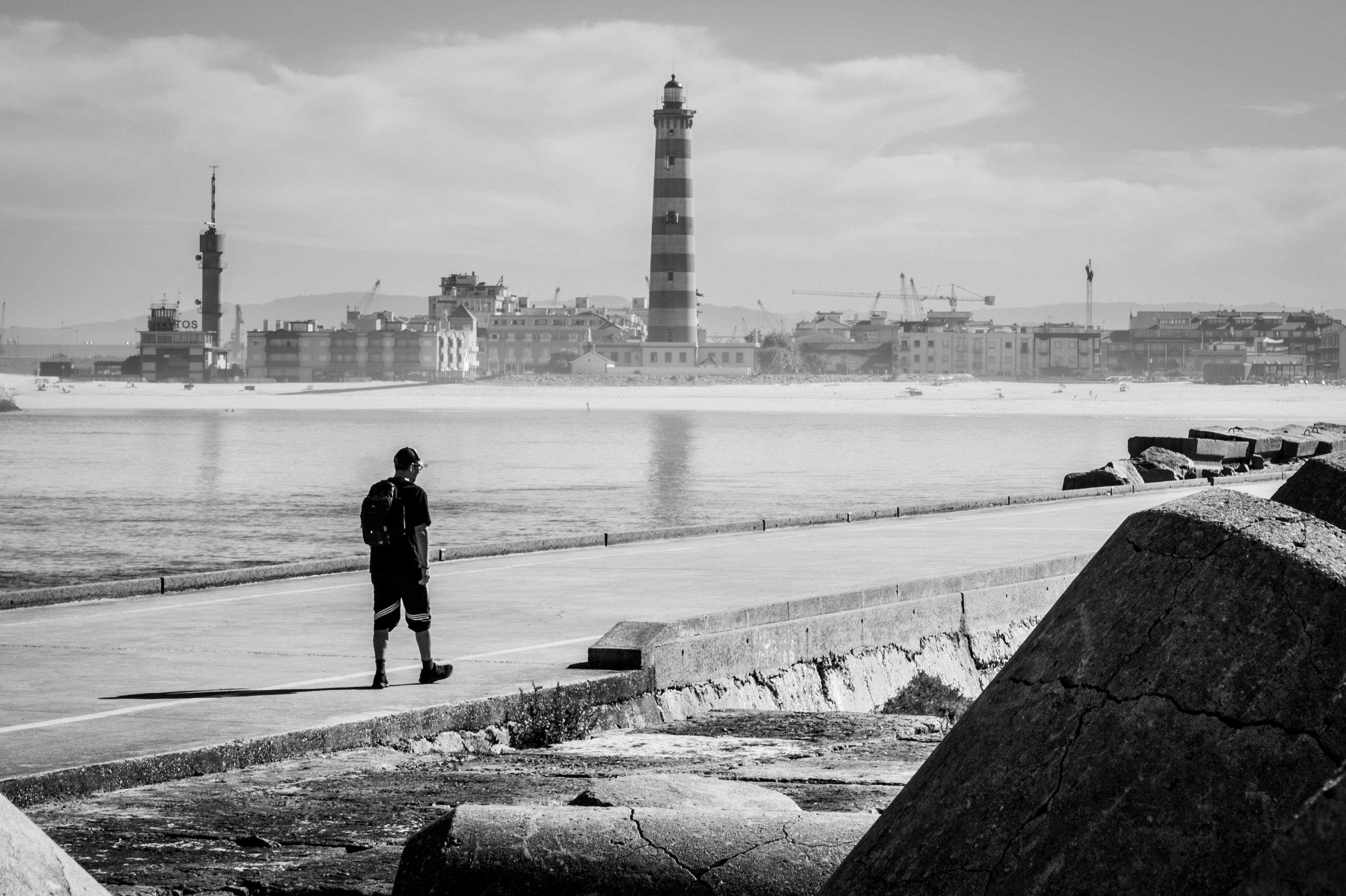 一张黑白照片，展现了一个宁静的场景：沿海的灯塔孤独地矗立着，而一位步行者独自行走在海边