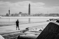 一张黑白照片，展现了一个宁静的场景：沿海的灯塔孤独地矗立着，而一位步行者独自行走在海边