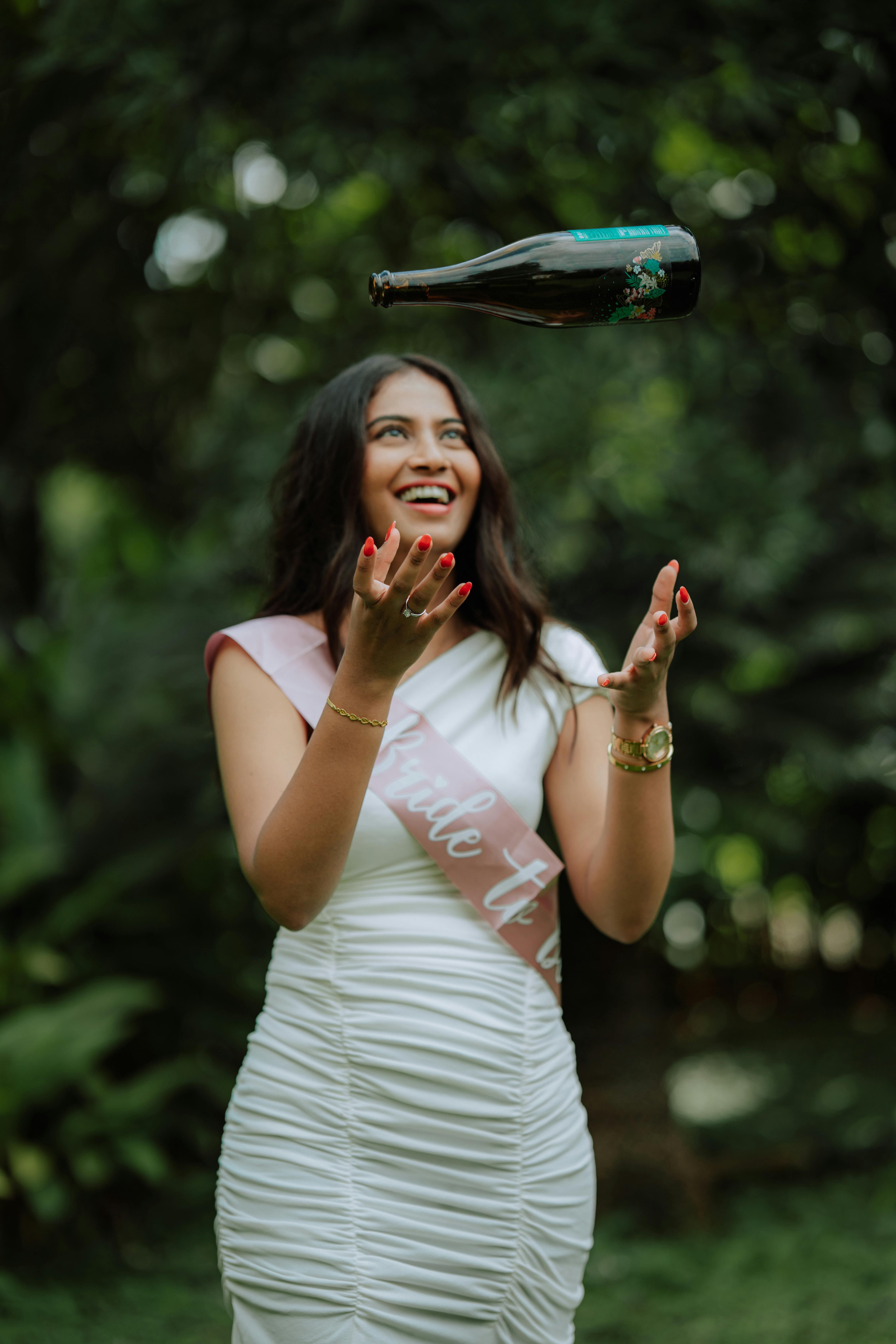 Bride-to-be wearing sash tosses champagne bottle in outdoor bridal shower.