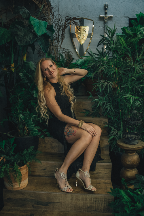 Free Woman in a black dress seated on stairs surrounded by plants, exuding elegance. Stock Photo