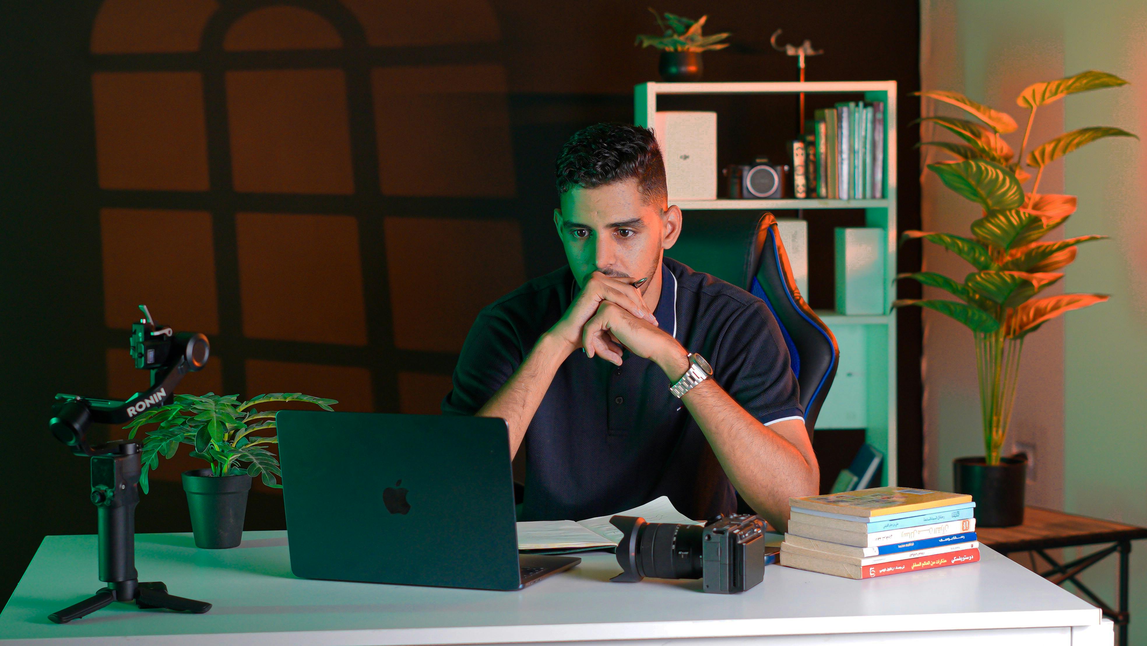 Man at desk with laptop, camera, books, and plants. Cozy workspace.