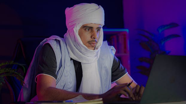 Middle Eastern man in traditional attire working intently on a laptop indoors at night.