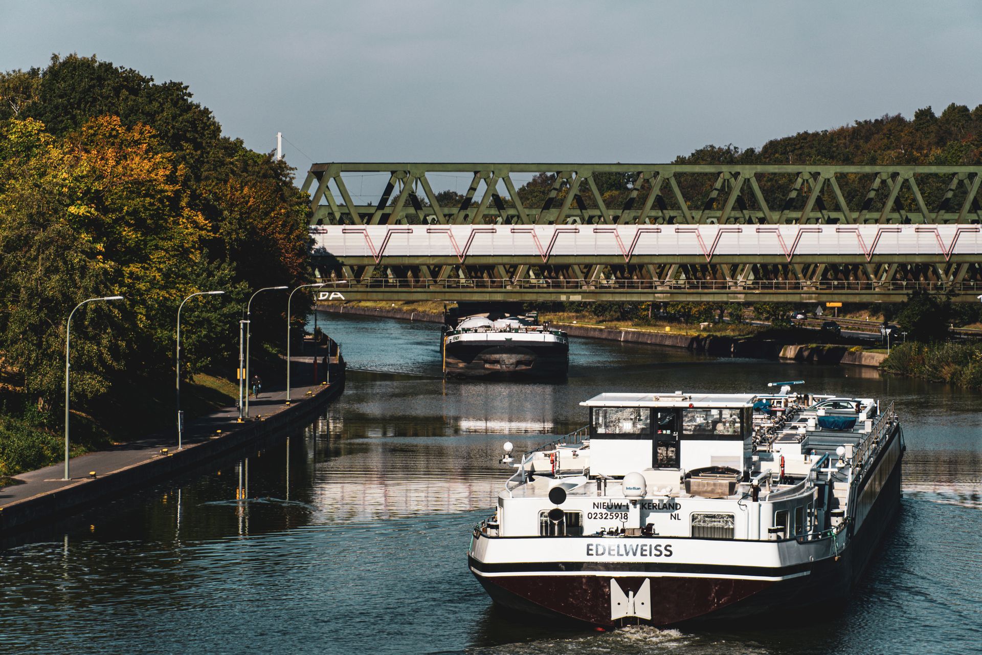 Large river barge navigating a scenic European canal beneath an iron bridge.