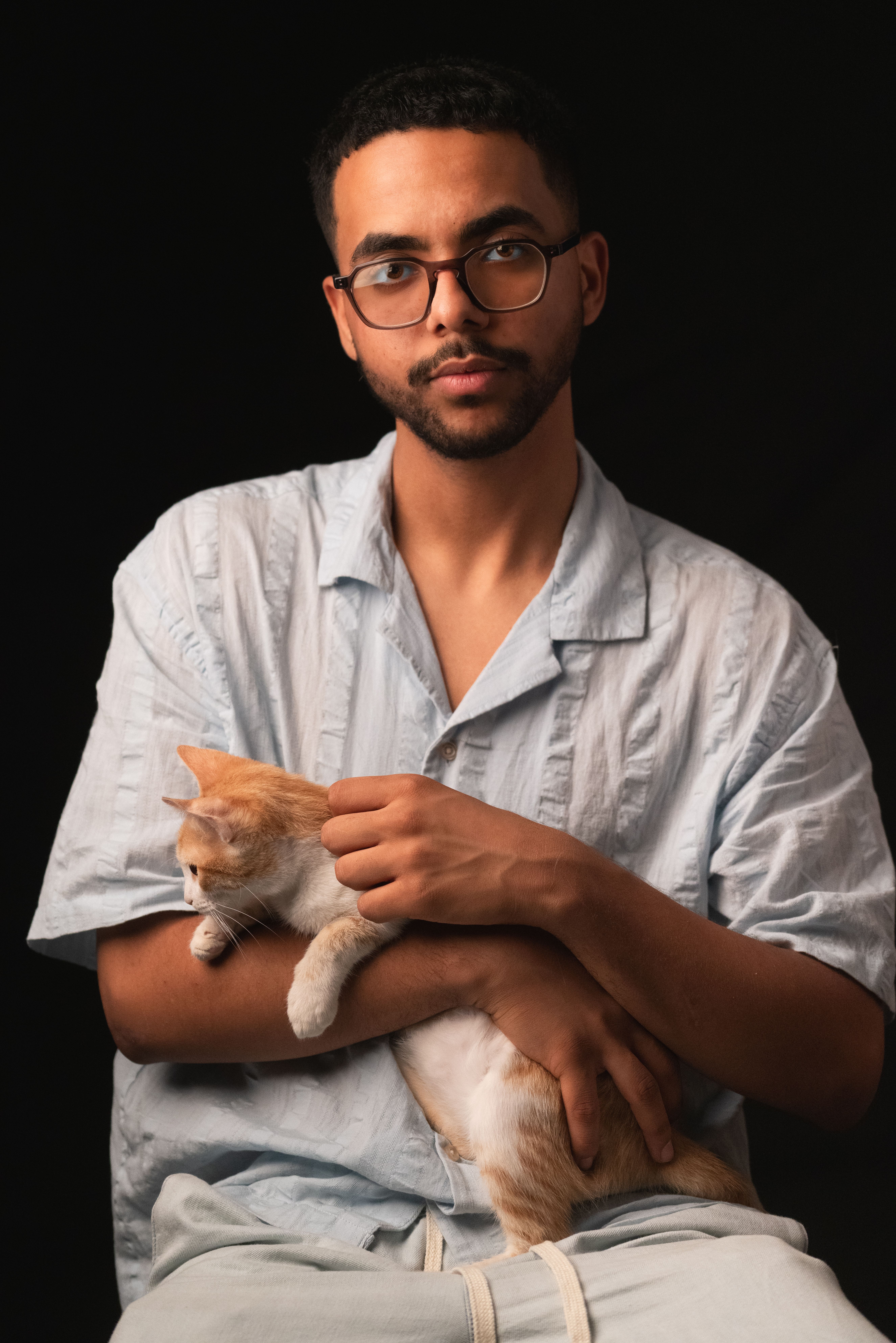 Portrait of a young man with glasses holding a cat against a dark background, showcasing a serene bond.
