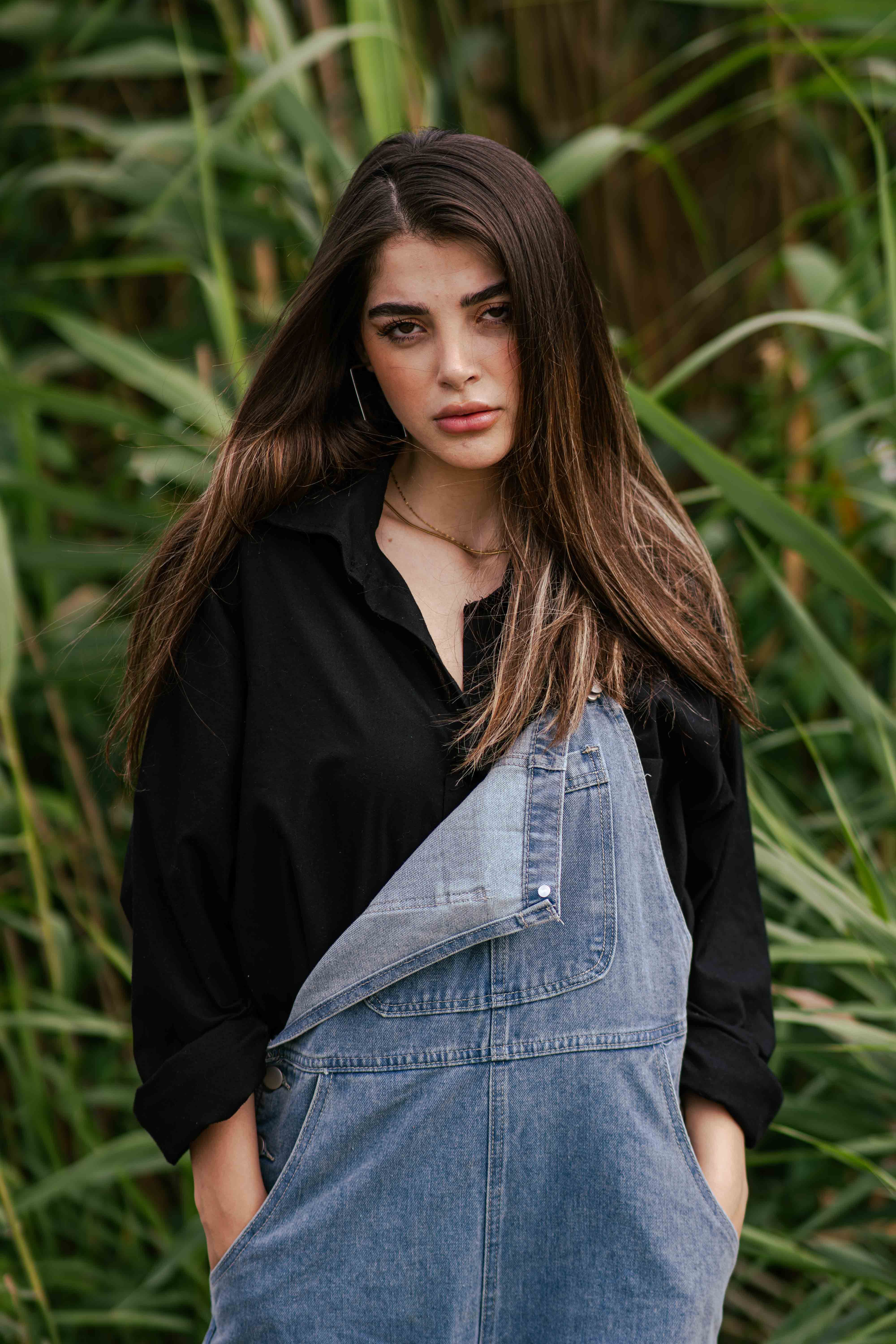 Portrait of a young woman in overalls standing outdoors surrounded by lush greenery.